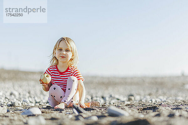 Cute blond girl holding apple sitting at beach on sunny day