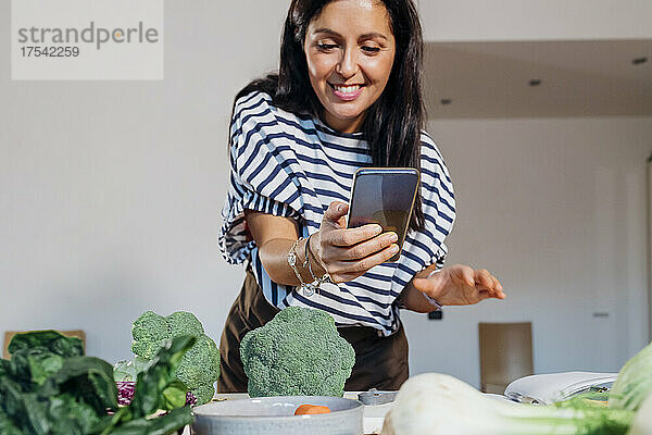 Lächelnde Frau  die zu Hause frisches Gemüse auf dem Tisch fotografiert