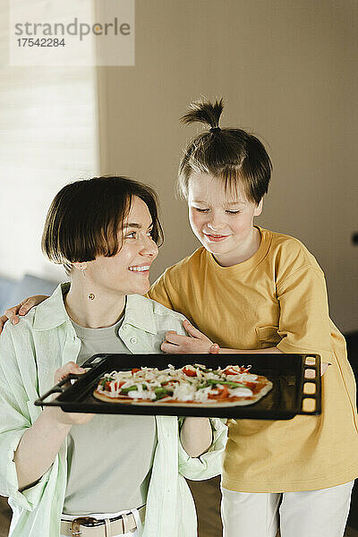 Lächelnde Mutter hält Pizzateig im Tablett und schaut ihren Sohn mit Pferdeschwanz an