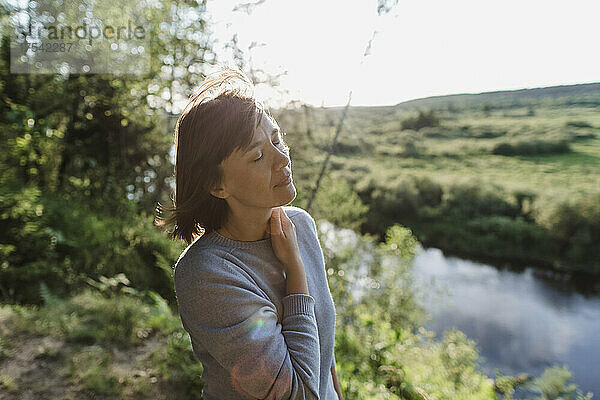 Frau mit geschlossenen Augen steht an einem sonnigen Tag am Flussufer