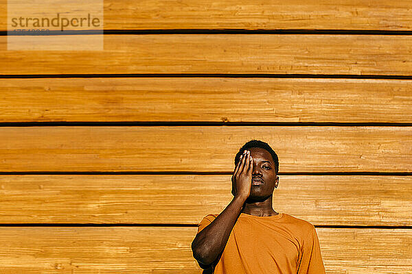 Man with hand covering eye in front of orange wall at sunset