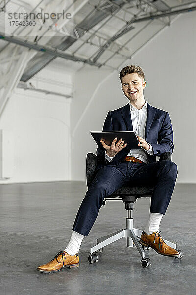 Lächelnder Geschäftsmann hält Tablet-PC auf Stuhl in der Industriehalle
