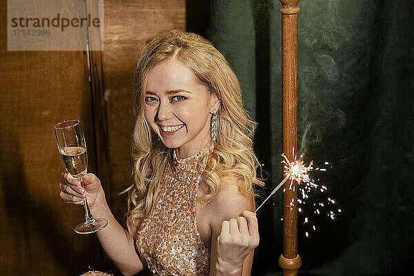 Cheerful young blond woman holding champagne flute and sparkler at New Year's party