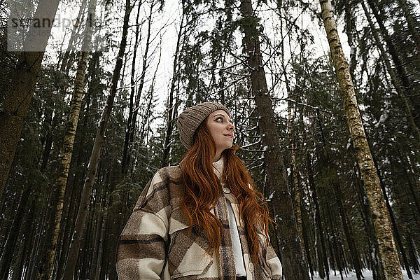 Nachdenkliche rothaarige Frau mit Strickmütze im Winterwald