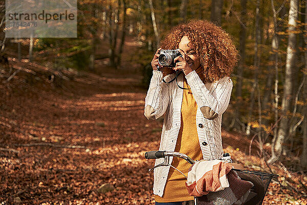 Woman with bicycle photographing through camera in autumn forest