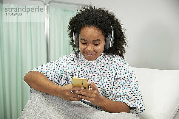 Smiling patient with headphones using smart phone sitting on bed at hospital