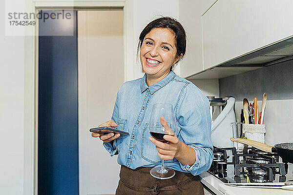 Glückliche Frau mit Smartphone und Weinglas in der heimischen Küche