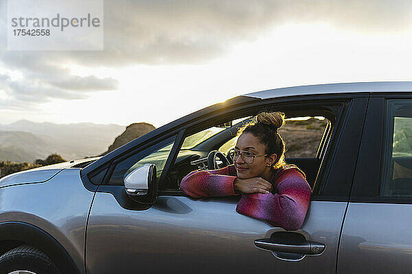 Lächelnde Frau lehnt sich bei Sonnenuntergang aus dem Autofenster