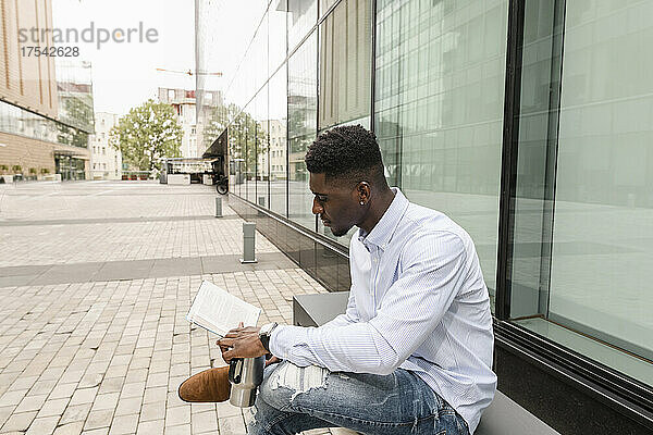Young man with coffee cup reading book in front of glass building
