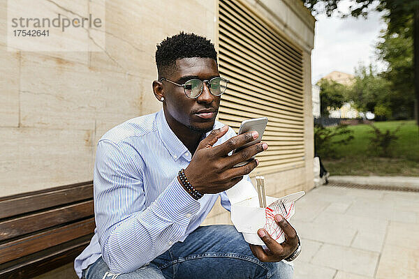Junger Mann mit Lunchbox und Smartphone auf Bank