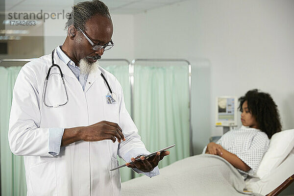 Male doctor using tablet PC with patient on bed in background at hospital
