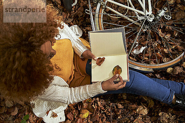 Woman with bicycle keeping leaf inside book sitting at autumn forest