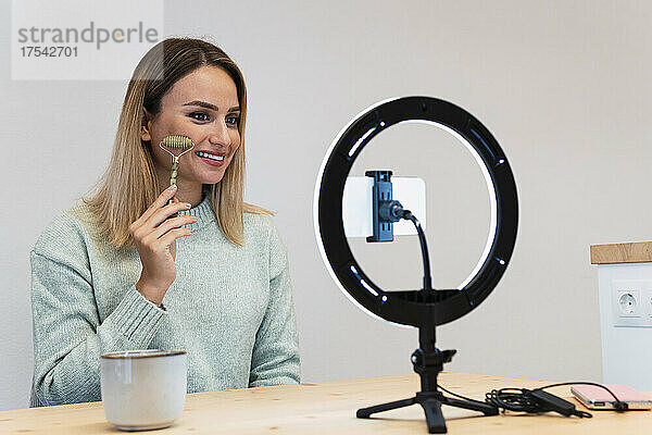 Smiling young beautician making tutorial with jade roller in front of ring light