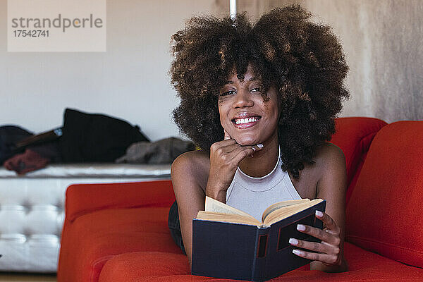 Smiling woman with book lying on sofa
