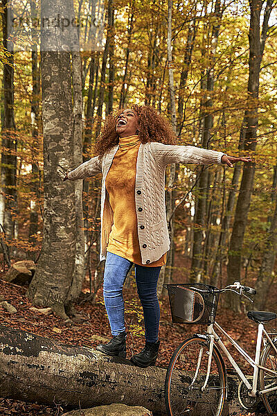 Happy woman with arms outstretched walking on log by bicycle in autumn forest
