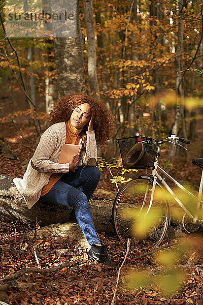 Woman with hand in hair holding book sitting on log by bicycle in autumn forest