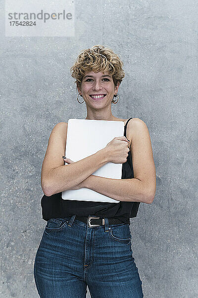Lächelnde junge Geschäftsfrau hält Laptop vor grauer Wand