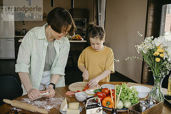 Smiling mother looking at son cutting tomato on table