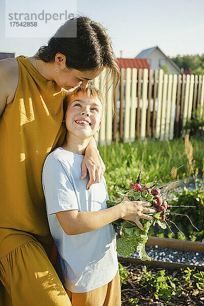 Happy mother embracing smiling son holding radish at garden