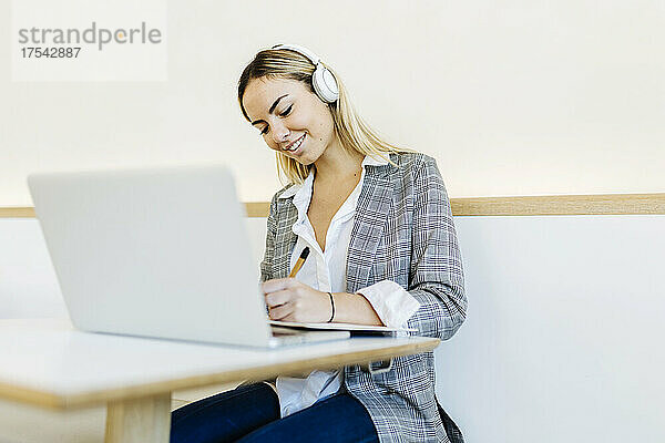 Smiling freelancer with headphones writing on book in cafe