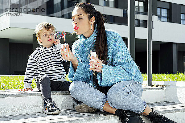 Playful mother blowing soap bubble by son in city
