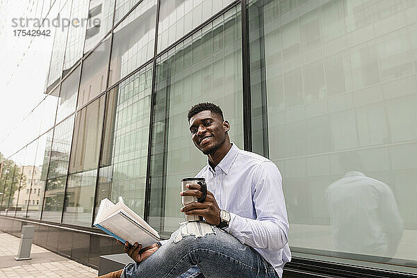 Smiling man with book and coffee cup in front of building