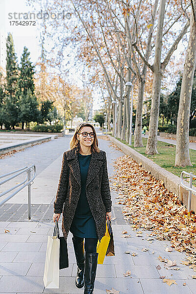 Lächelnde Frau mit Einkaufstüten  die im Herbst auf dem Fußweg spaziert
