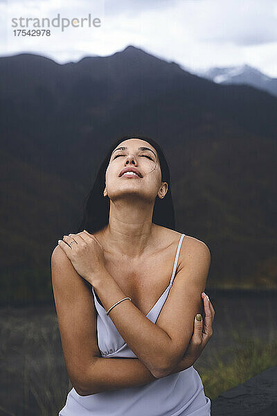 Sensuous young woman with eyes closed hugging herself in front of mountain
