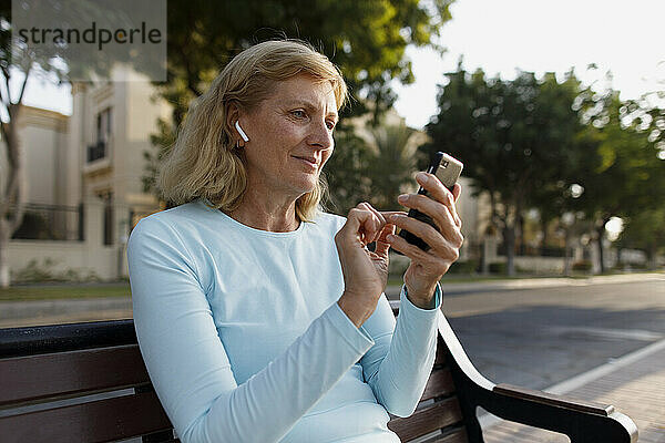 Ältere Frau mit In-Ear-Kopfhörern und Smartphone auf Bank sitzend