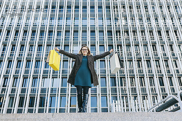 Cheerful woman holding shopping bags standing in front of building in city
