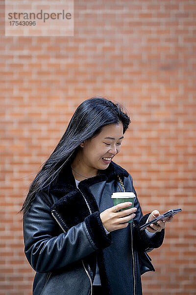 Smiling woman with disposable cup using mobile phone in front of wall