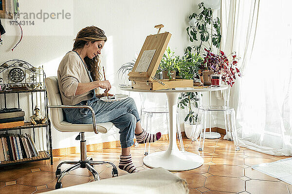 Künstler mischt Farben in einer Palette und sitzt zu Hause auf einem Stuhl vor der Staffelei