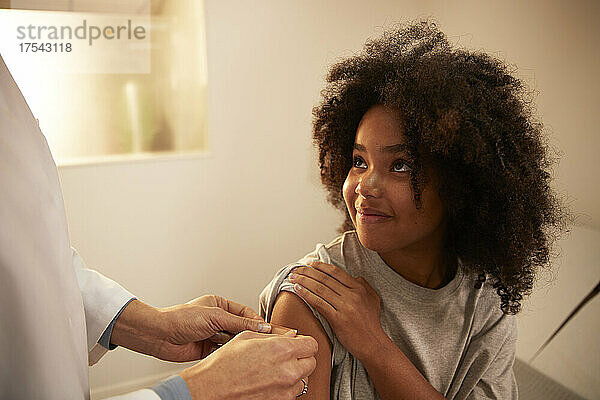 Lächelndes Mädchen schaut dem Arzt zu  der im Krankenhaus einen Klebeverband am Arm anlegt