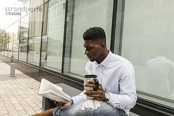 Junger Mann mit wiederverwendbarer Kaffeetasse liest ein Buch vor dem Gebäude