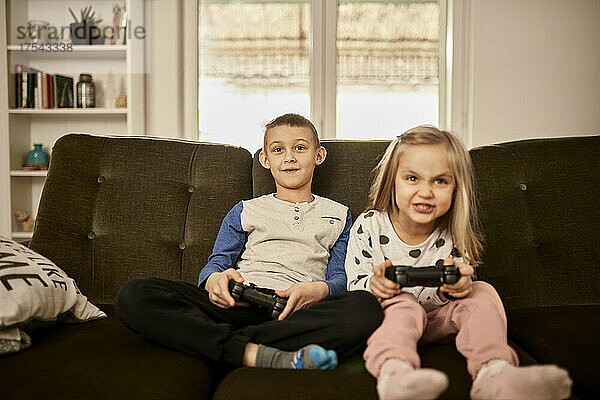 Brother and sister playing video game sitting on sofa at home