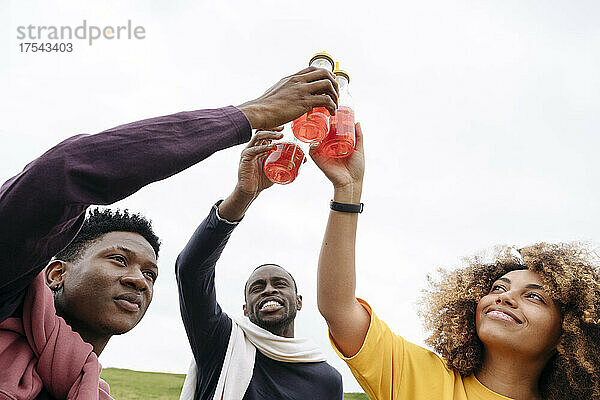 Smiling friends toasting lemonade bottles in front of clear sky