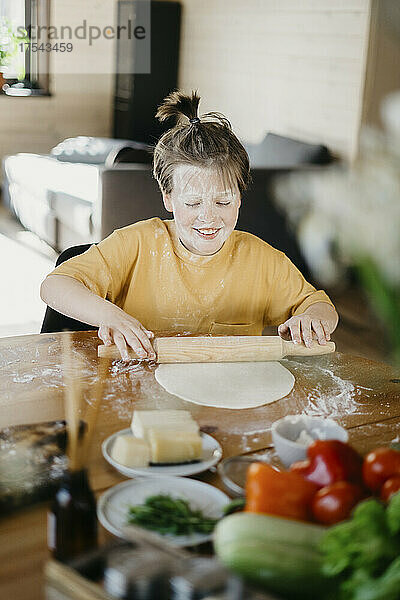Smiling boy with flour on face rolling pin on dough at home
