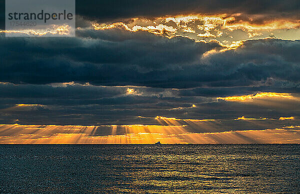 Sonnenstrahlen und goldene Wolken über dem Meer bei Sonnenaufgang mit einem kleinen Fischerboot in der Ferne