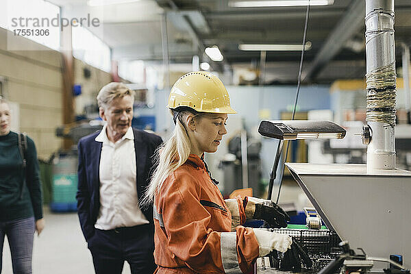 Weiblicher Industriearbeiter mit Schutzhelm bei der Arbeit an Maschinen  während im Hintergrund Manager in einer Fabrik zu sehen sind