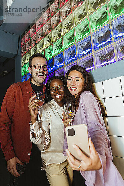 Fröhliche Frau nimmt Selfie mit männlichen und weiblichen Freunden durch Smartphone in Bar