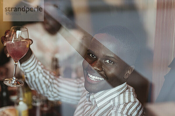 Porträt eines lächelnden jungen Mannes mit Weinglas  gesehen durch das Glas eines Restaurants