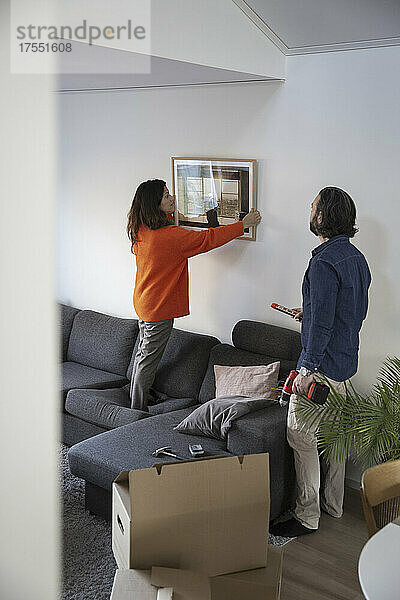 Frau hängt Bilderrahmen an Wand von Mann im Wohnzimmer
