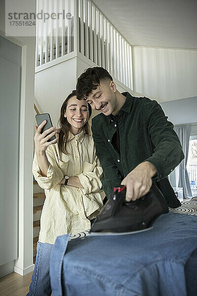 Lächelnder Mann  der sein Hemd bügelt  während seine Freundin mit ihrem Smartphone zu Hause steht
