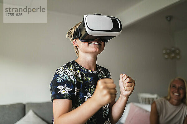 Lächelnder Junge  der ein Virtual-Reality-Headset trägt  während seine Mutter zu Hause im Hintergrund sitzt