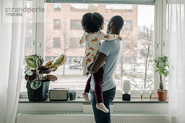 Vater  der seine Tochter trägt  während er durch das Fenster seines Hauses schaut