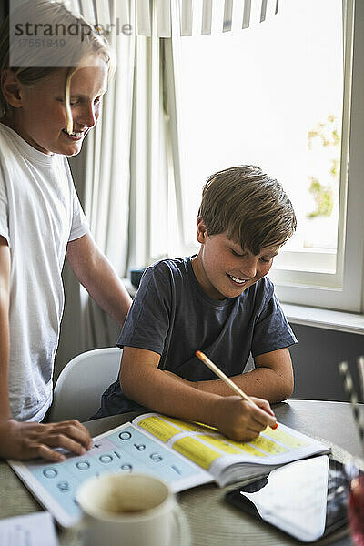 Lächelnder Junge neben seinem Bruder  der zu Hause Hausaufgaben macht