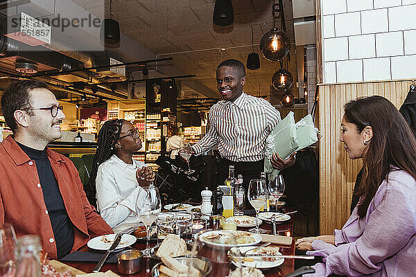 Glücklicher Mann im Gespräch mit männlichen und weiblichen Freunden während einer Dinnerparty im Restaurant