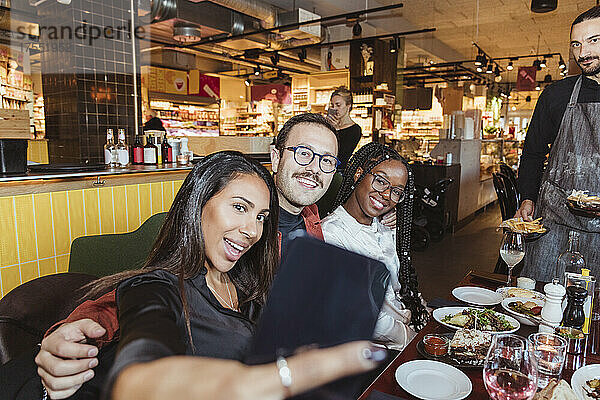 Fröhliche Frau  die ein Selfie über ihr Smartphone mit männlichen und weiblichen Freunden im Restaurant macht