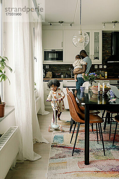 Vater trägt kleinen Jungen  während seine Tochter in der Küche Roller fährt