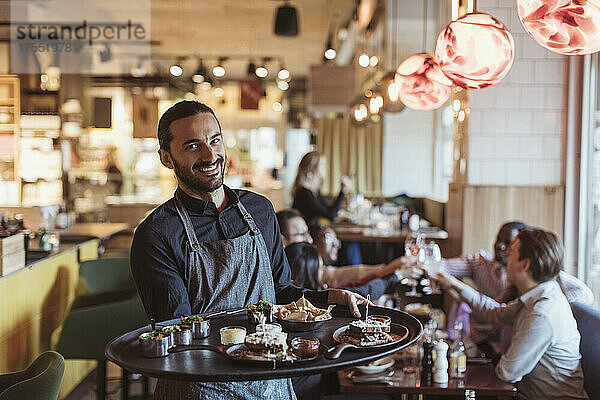Porträt eines männlichen Kellners mit einem Essenstablett  während die Kunden im Hintergrund an der Bar anstoßen